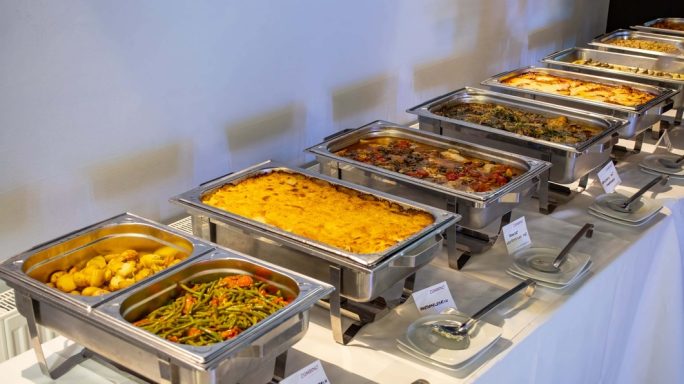 Buffet mit verschiedenen Gerichten in Warmhaltetheken auf weißem Tisch.