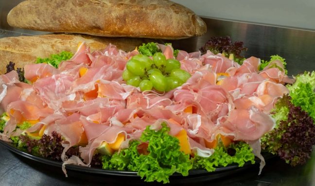 Ein Teller mit Schinken, green Trauben und buntem Salat auf einem Tisch.
